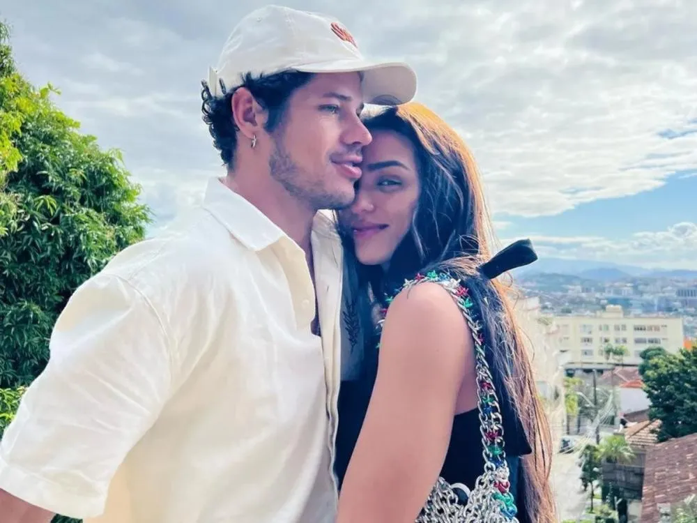 José Loreto, usando boné e camisa branca, abraça carinhosamente Rafa Kalimann, que veste blusa escura com detalhes coloridos. O casal está ao ar livre, com paisagem urbana ao fundo. Imagem representa o relacionamento de José Loreto e Rafa Kalimann.